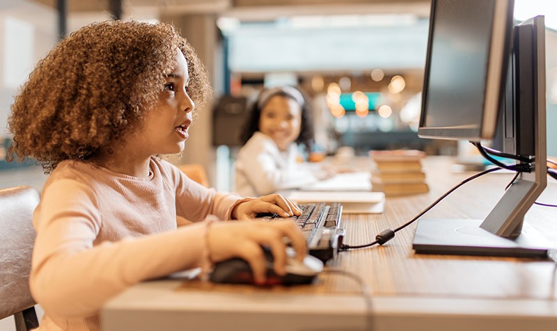 Foto: Een kind werkt geconcentreerd op de computer in een bibliotheek, met stapels boeken op de achtergrond.