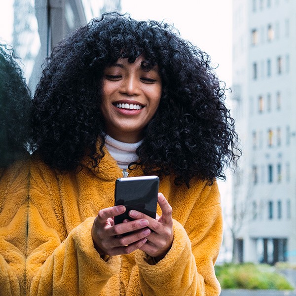 inset_wise_nl_4 Een persoon met een gele jas staat buiten en houdt een mobiele telefoon vast. Op de achtergrond zijn gebouwen en een glazen reflectie zichtbaar.
