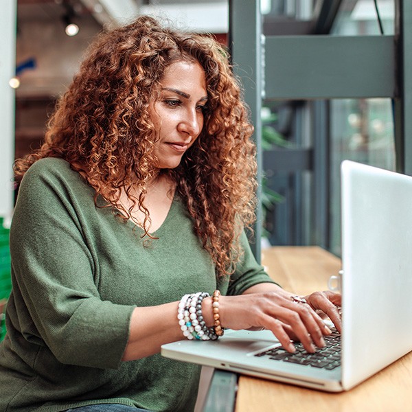inset_wise_nl_2 Een persoon met krullend haar zit aan een houten tafel en typt op een laptop. Op de achtergrond is een lichte ruimte met grote ramen zichtbaar.