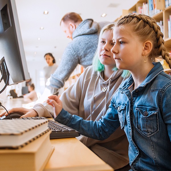 inset_wise_nl_1 Een jong meisje in een spijkerjasje zit naast een andere persoon achter een computer in een bibliotheek. Op de voorgrond liggen boeken, en op de achtergrond zijn andere bezoekers en boekenplanken zichtbaar.