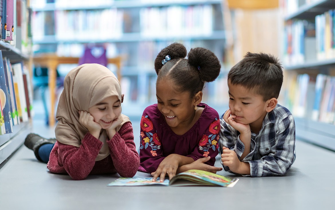 Drie kinderen lezen samen een boek op de vloer van een bibliotheek, omringd door boekenkasten.